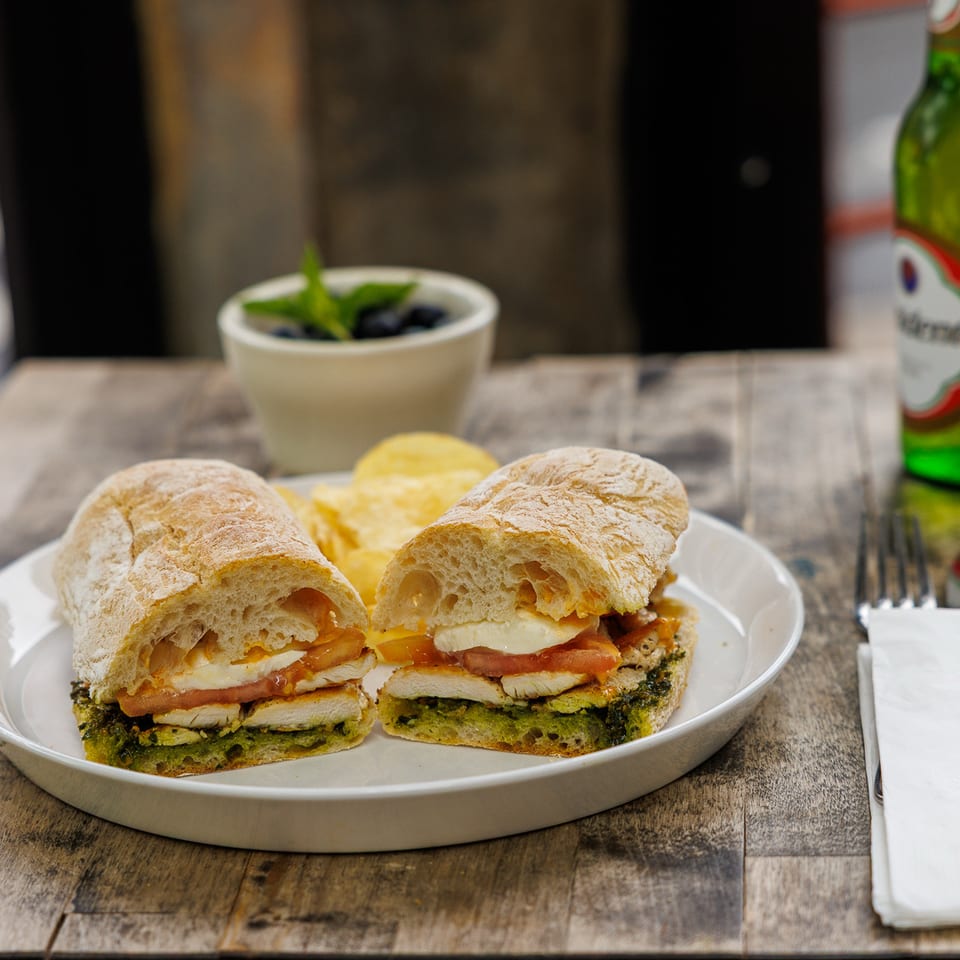 Plate of two chicken sandwiches with tomato, cheese, and greens, served with potato chips, dip, and a green beer bottle on a rustic table.