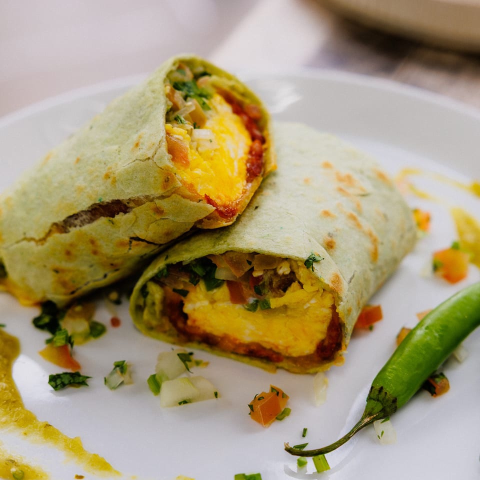 Two green tortilla burritos filled with melted cheese, vegetables, and salsa, served with whole green chilies on a white plate.