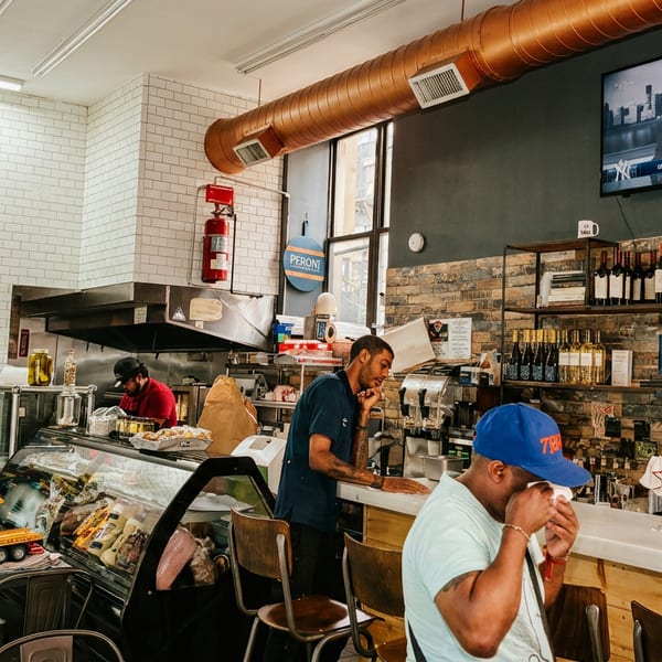 Customers and staff inside a bustling deli with a display case, bar seating, and a TV showing news.