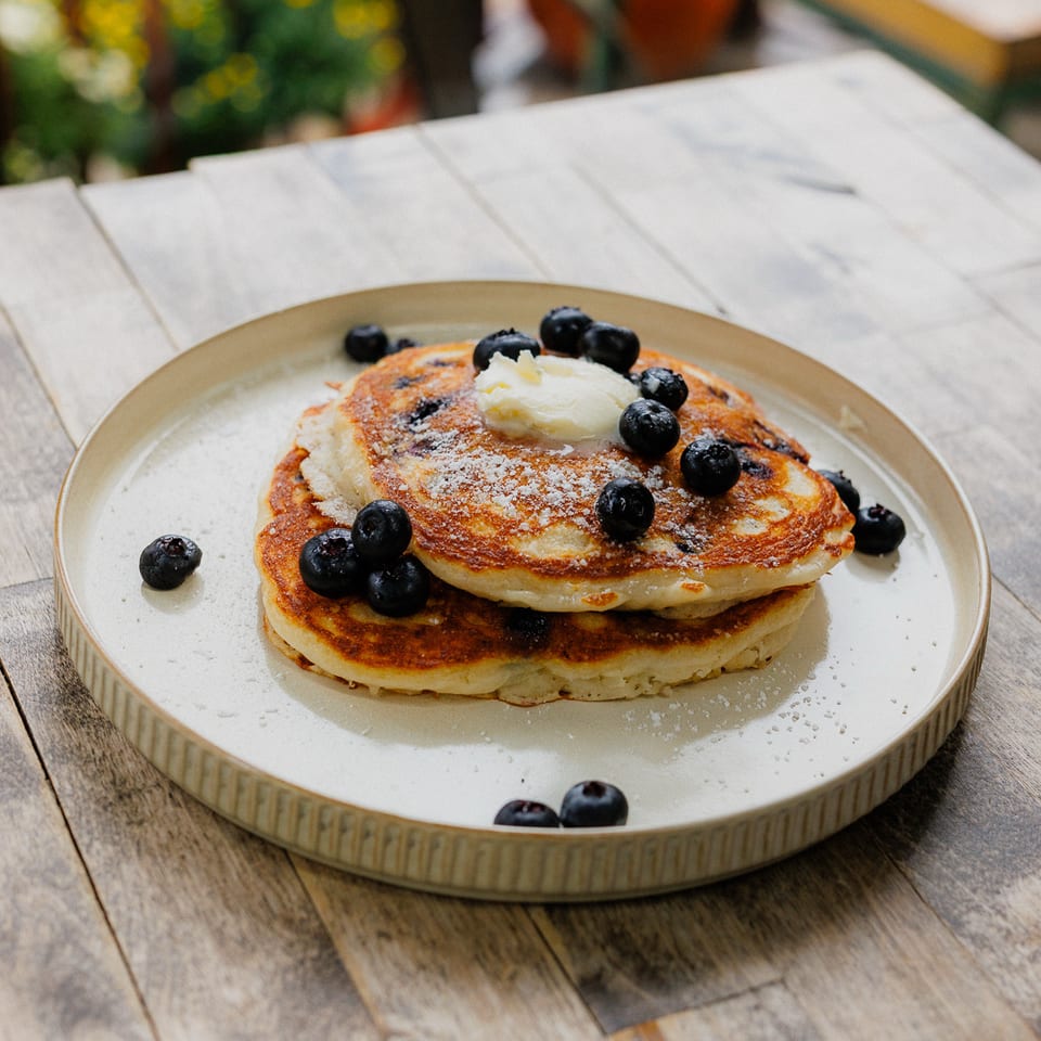 Stack of golden pancakes topped with butter, blueberries, and powdered sugar on a plate at an outdoor table.