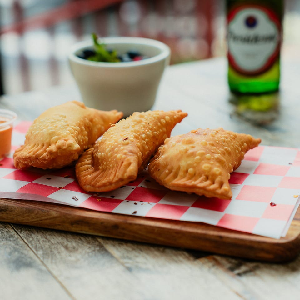 Three golden fried empanadas sit on a checkered paper-lined wooden board with dipping sauce, salad, and a beer bottle.