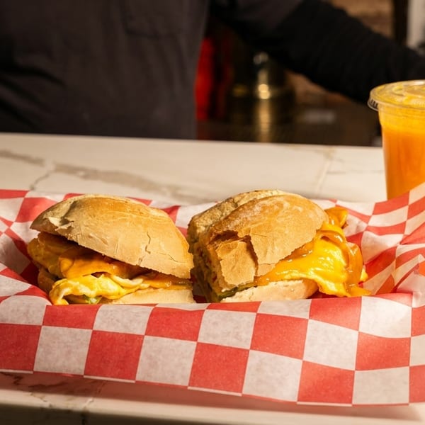 Two breakfast sandwiches with melted cheese in a red and white checkered basket, with an orange drink.
