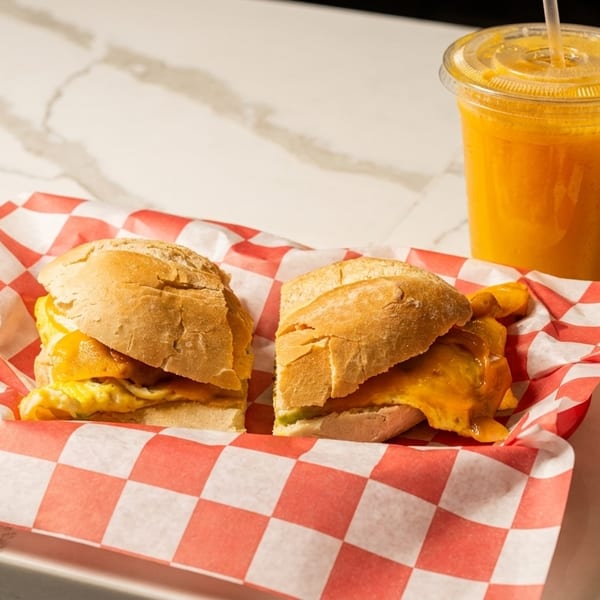 Two breakfast sandwiches with melted cheese and scrambled eggs in a red checkered basket with an orange juice.