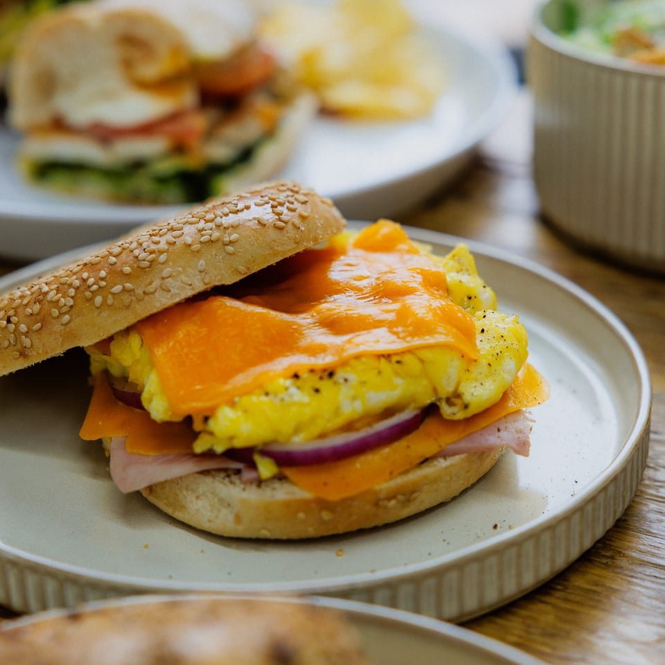 Sesame bagel sandwich layered with scrambled eggs, melted cheese, ham, and red onion on a plate.