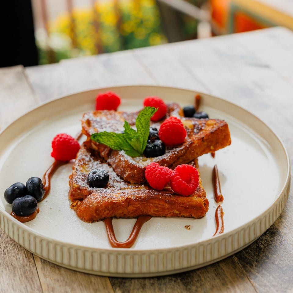 French toast topped with powdered sugar, raspberries, blueberries, mint, and chocolate sauce on a ceramic plate.